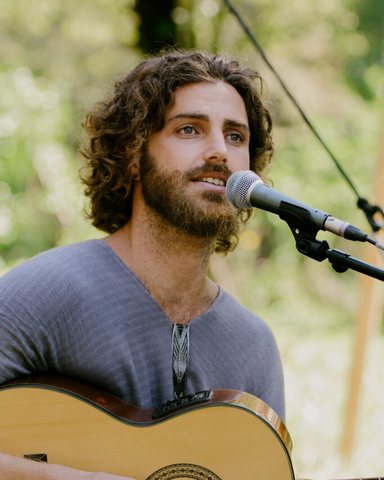 Natan Rabin singing into a microphone and playing an acoustic guitar outdoors, trees soft behind him.