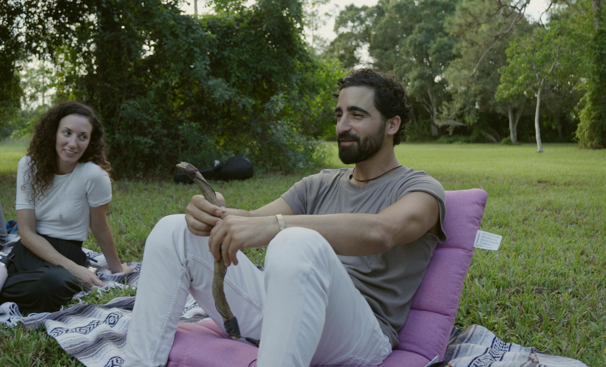 Two people sitting and talking on a blanket on the grass, trees behind them.